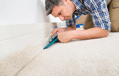 A carpet fitter in a blue shirt cutting a cream carpet with a high quality underlay to size with a Stanley knife